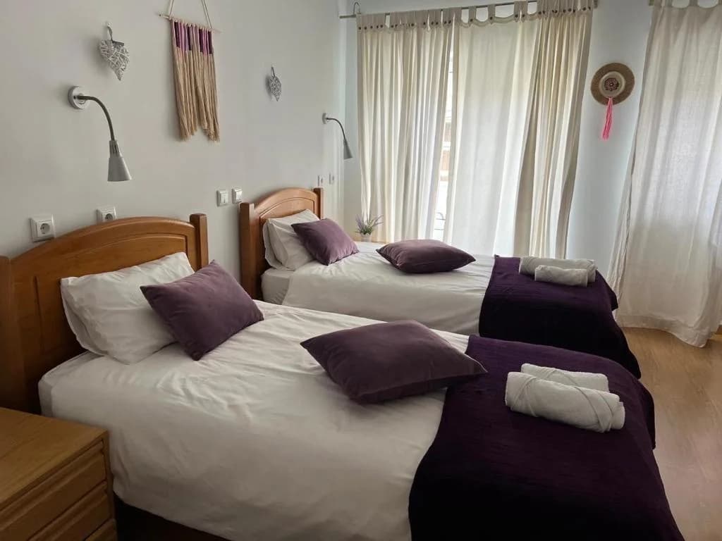 Quarto Ametista na Casa Miná com duas camas individuais, roupa de cama branca e detalhes roxos, luz natural e cortinas claras