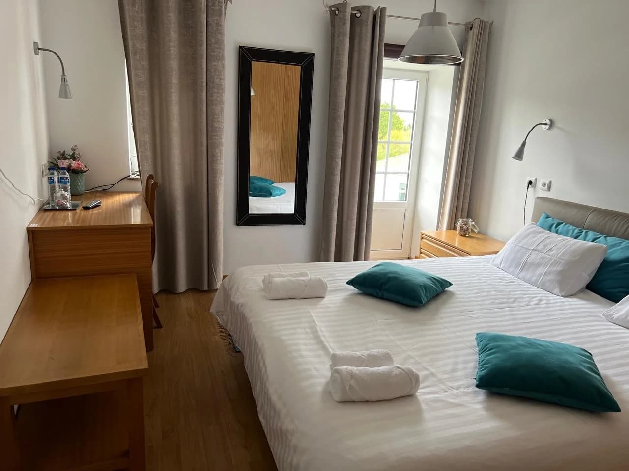 Quarto luminoso com cama de casal, roupa de cama branca, almofadas em tons azuis, mesa de madeira e janela com cortinas claras.